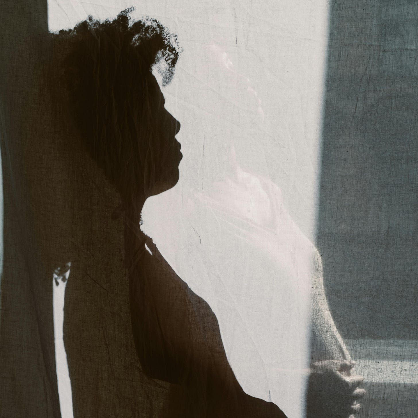 Silhouette of a woman with curly hair, standing against a softly lit fabric backdrop, creating a serene and introspective atmosphere.