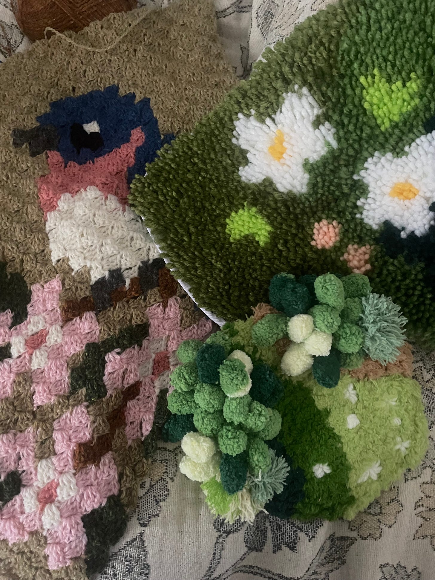 an image of three projects in various states of completion. One is a crochet project depicting a bluebird on a cherry blossom branch; another is an latch hook rug depicting daisies and grasses; the final project is a mossy coaster made with pom pom, fluffy yarns and embroidered flowers