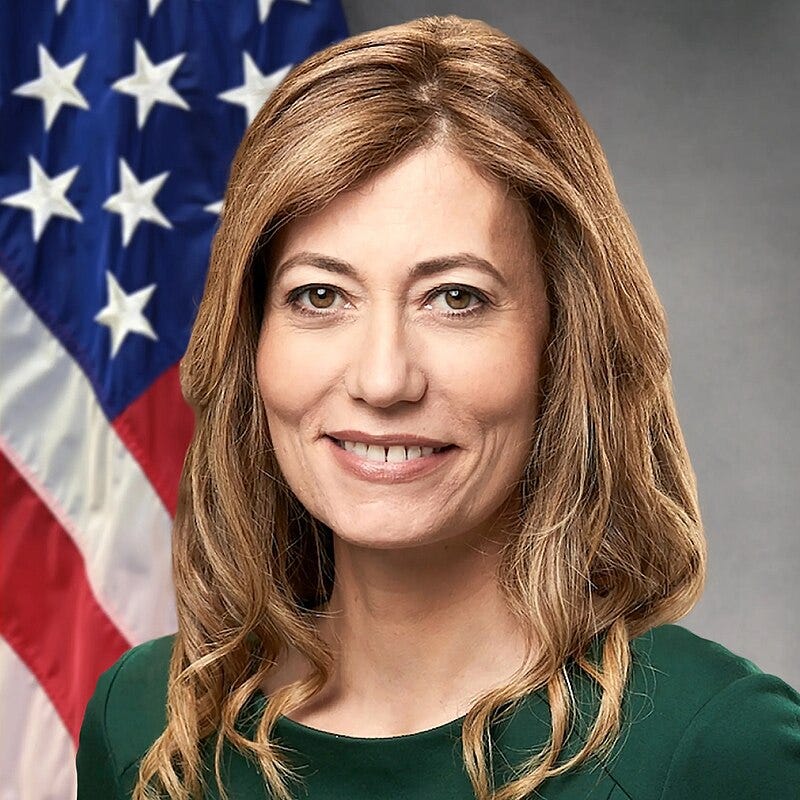 A picture of a smiling women, facing the camera, in front of a gray background and the U.S. flag