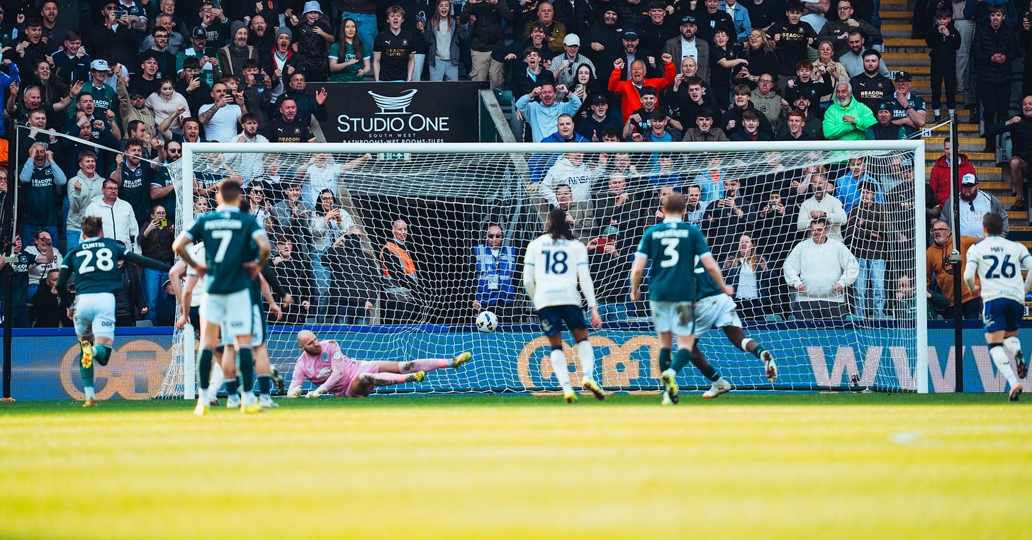 A Plymouth Argyle player scores past a diving Huddersfield Town goalkeeper, with the Plymouth end behind the goal erupting in celebration. A Plymouth Argyle player scores past a diving Huddersfield Town goalkeeper, with the Plymouth end behind the goal erupting in celebration.