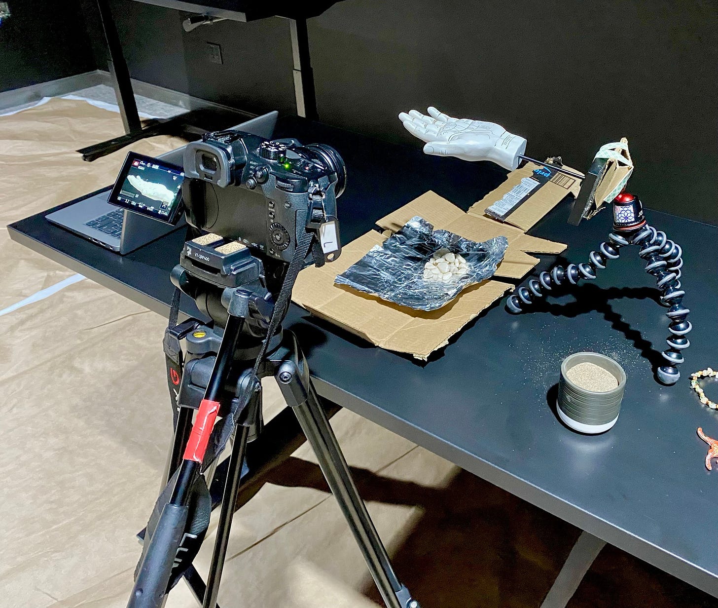 A DSLR camera on a tripod is pointed at a table, atop of which is a prop hand rigged to a flexible mount above cardboard and foil. A small pot of sand and a laptop are on the table as well.