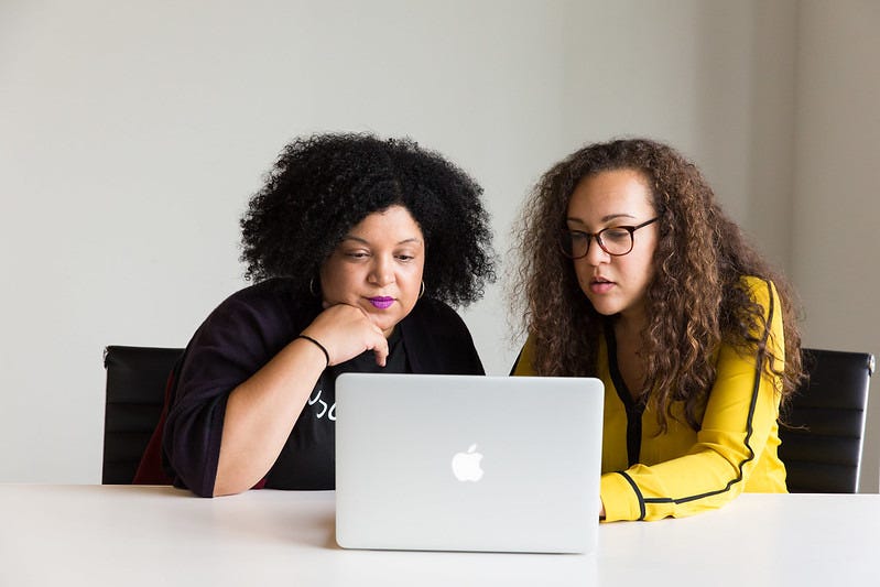 Two people sit next to each other, looking intently at a laptop. Both have thick, curly, fairly long hair. Two people sit next to each other, looking intently at a laptop. Both have thick, curly, fairly long hair.