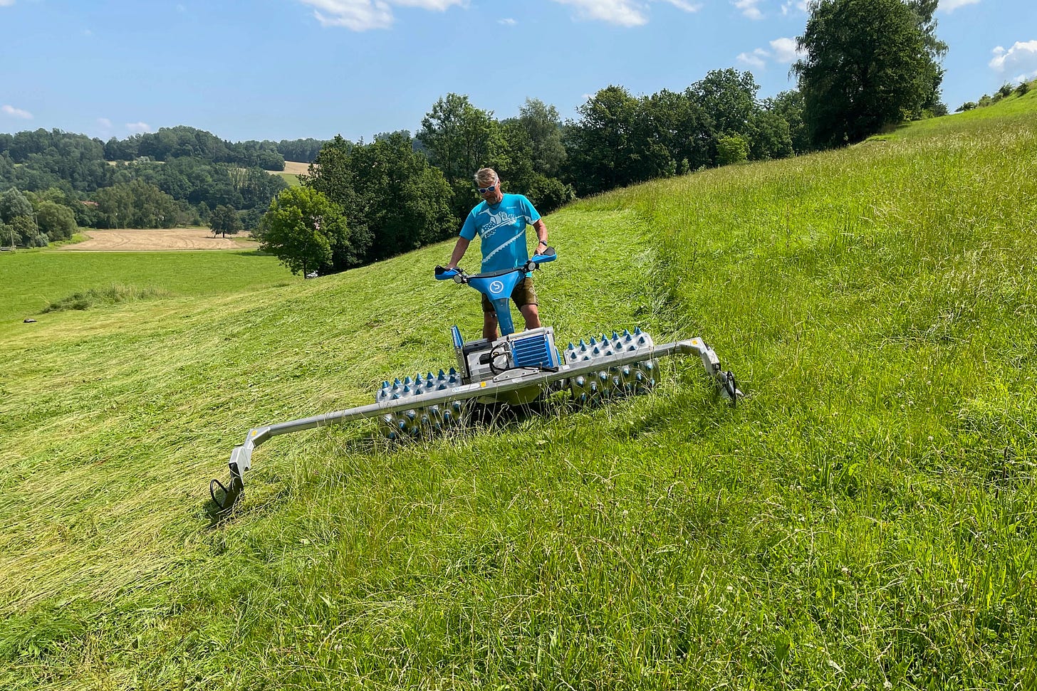 Ein Landwirt mäht an einem sonnigen Tag eine steile, grüne Wiese mit einem blauen Brielmaier Motormäher. Der Mann trägt ein türkisfarbenes T-Shirt, Sonnenbrille und kurze Hosen und steuert die Maschine mit beiden Händen. Der Motormäher fährt auf breiten Alu-Stachelwalzen, ideal für den Einsatz an Hanglagen. Im Hintergrund sind sanfte Hügel, Felder, Bäume und ein blauer Himmel mit wenigen Wolken zu sehen. Ein Landwirt mäht an einem sonnigen Tag eine steile, grüne Wiese mit einem blauen Brielmaier Motormäher. Der Mann trägt ein türkisfarbenes T-Shirt, Sonnenbrille und kurze Hosen und steuert die Maschine mit beiden Händen. Der Motormäher fährt auf breiten Alu-Stachelwalzen, ideal für den Einsatz an Hanglagen. Im Hintergrund sind sanfte Hügel, Felder, Bäume und ein blauer Himmel mit wenigen Wolken zu sehen.