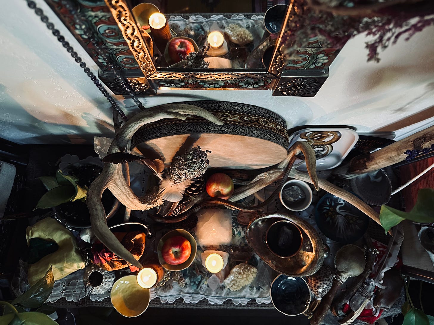 Above view of Pam's autumnal equinox altar. In view is a smallish, ornate mirror reflecting the altar with prayer beads hanging in front. On the altar is a rain drum leaning up against the wall and many, many items including two apples, deer antlers, bird feathers, pine cones, crystals, a copper bowl of water in an abalone shell, dried lavender, a rattle and other sacred tools, two candles and a small yellow bowl for day and a small blue bowl for night.