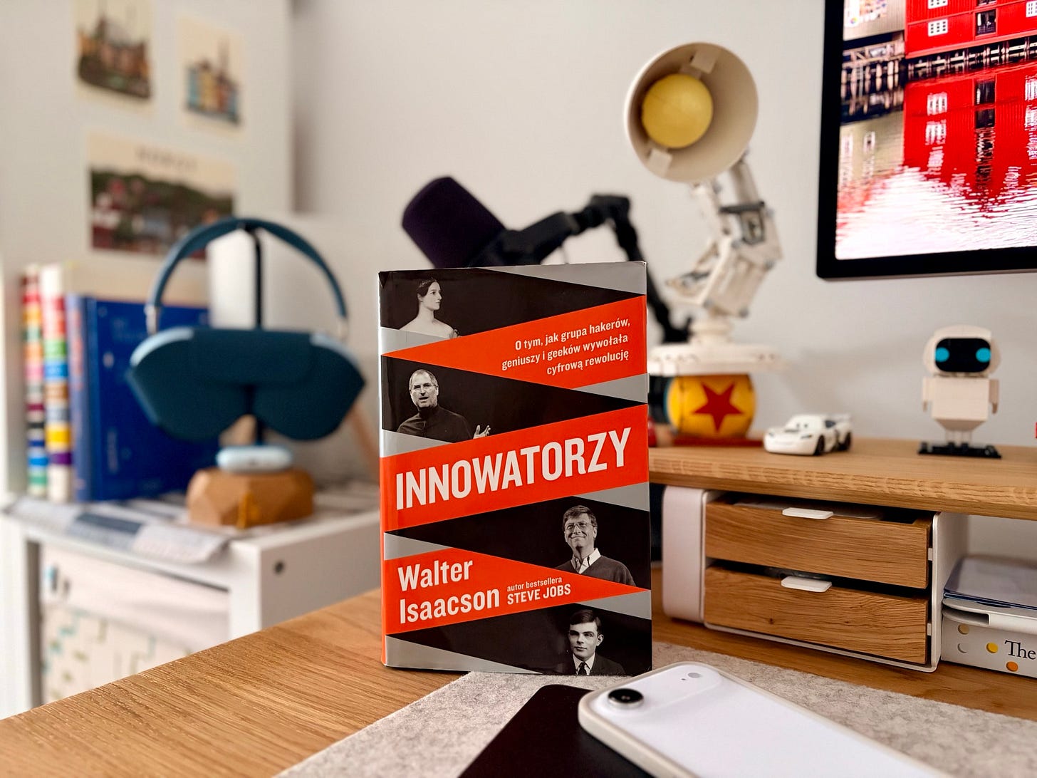 Na zdjęciu książka „Innowatorzy” stojąca na drewnianym biurku, w otoczeniu współczesnych gadżetów