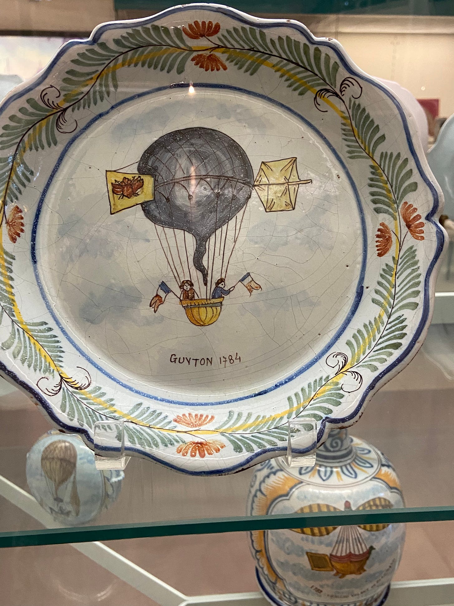 A decorative 18th-century plate featuring two balloonists waving French flags, held aloft by a hot air balloon