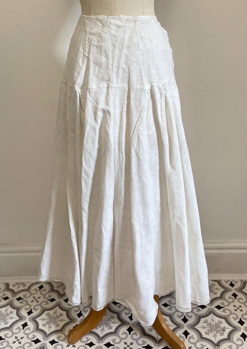 May include: A long, white skirt made of a lightweight fabric, likely linen or cotton, is displayed on a mannequin. The skirt has a high waist and a full, flowing silhouette. The fabric appears slightly textured, with visible seams and pleats adding detail.