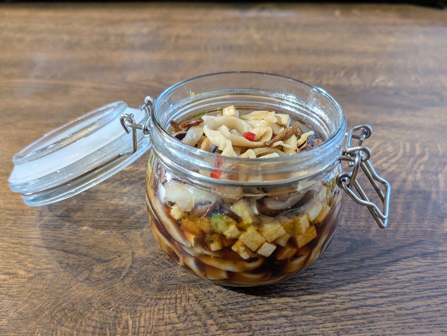 A glass jar with a metal clasp lid sits open on a wooden surface, filled with sliced mushrooms, tofu cubes, noodles, and small pieces of red chilli and green pepper, all submerged in a dark, glossy broth. The ingredients are tightly packed and colourful, giving the jar a rich, layered appearance.