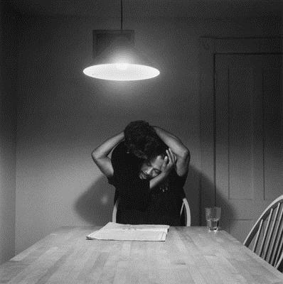 Foto em preto e branco. Um homem sentado numa cadeira na cabeceira de uma mesa é abraçado por trás por uma mulher em pé. Ele estica os braços para trás para abraçá-la também.  Em cima da mesa, há um jornal e um copo com algum líquido pela metade. O casal é iluminado por uma luminária pendente.