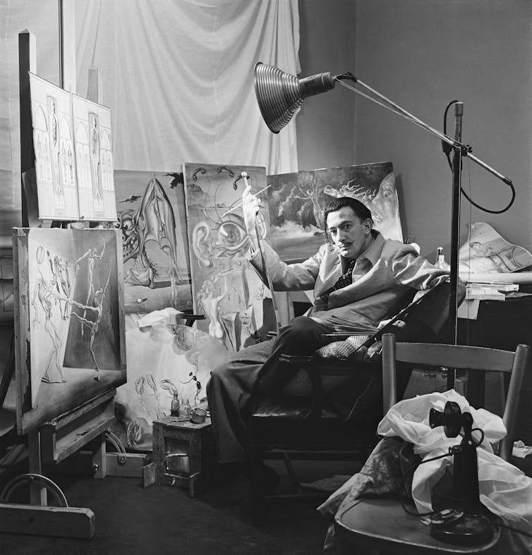 Black and white photo of man in suit sitting in chair in front of paintings on canvases