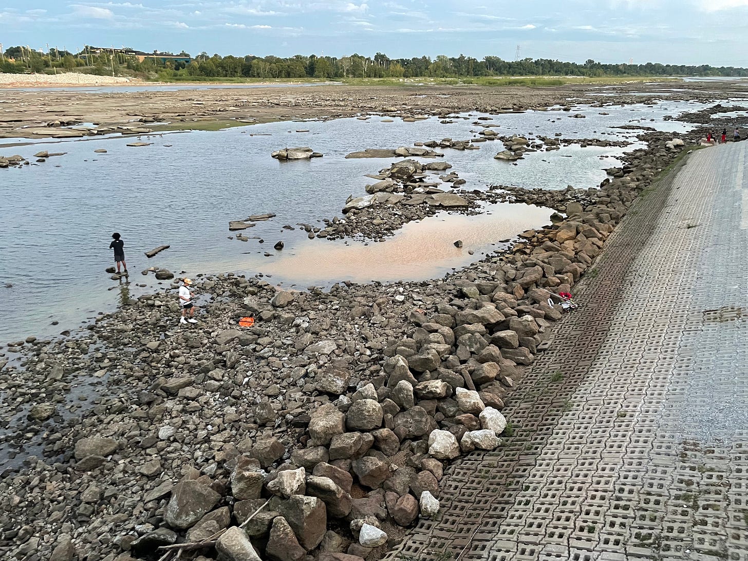 An articulated concrete embankment slopes down toward rocks where an opaque, rust colored plume is pooling and mixing with the Arkansas River. Five people and various items are visible among the rocks. One person is fishing on a rock in the area where the plume is mixing with the dark water of the channel. Another is walking among the rocks carrying a fishing pole. 