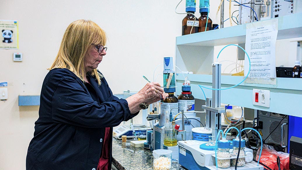 Pessoa usando óculos e casaco azul-marinho manipula um frasco de vidro em bancada de laboratório equipada com diversos frascos, tubos, equipamentos eletrônicos azuis na prateleira superior, tubo de silicone azul, computador, materiais diversos em recipientes brancos e azuis, com parede branca ao fundo e pôster à esquerda. Pessoa usando óculos e casaco azul-marinho manipula um frasco de vidro em bancada de laboratório equipada com diversos frascos, tubos, equipamentos eletrônicos azuis na prateleira superior, tubo de silicone azul, computador, materiais diversos em recipientes brancos e azuis, com parede branca ao fundo e pôster à esquerda.