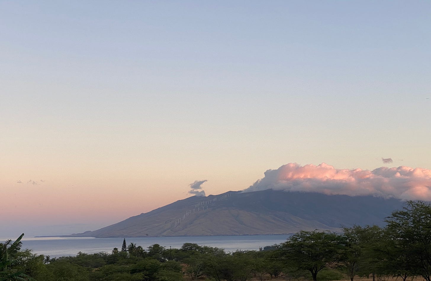 A faraway bay in dawn light next to a round mountain with a crown of pink-tinged clouds