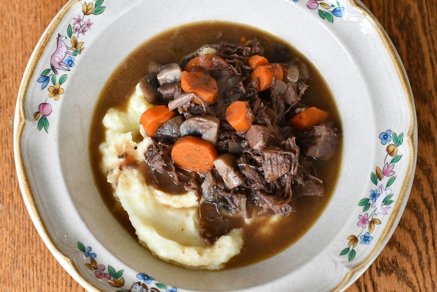 A bowl of boeuf bourguignon with chunks of beef, carrots and mushrooms in a dark gravy over mashed potatoes, in a cream bowl with a painted border of flowers and animals, on a wooden table.