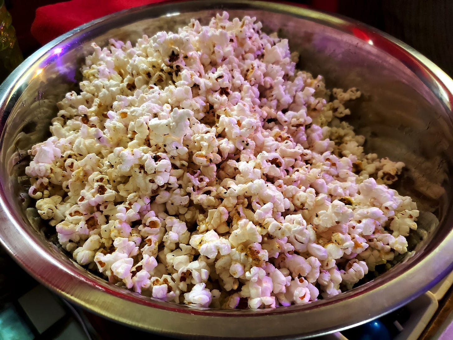 A large metal bowl is filled with freshly popped popcorn, showing fluffy kernels with a hint of seasoning. The vibrant colors reflect off the metal surface, adding a warm glow.