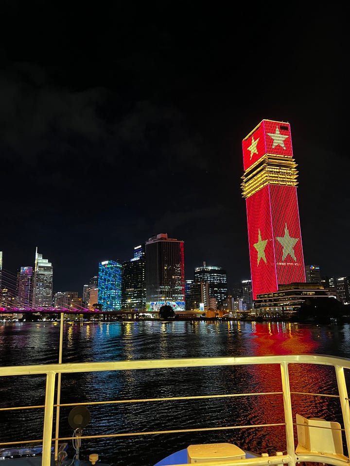 Nighttime view of Ho Chi Minh City skyline along the Saigon River, with illuminated high-rises—including a tower lit in red with yellow stars and another displaying “SAIGON”—and a glowing cable-stayed bridge, reflected in the water from a boat railing in the foreground.