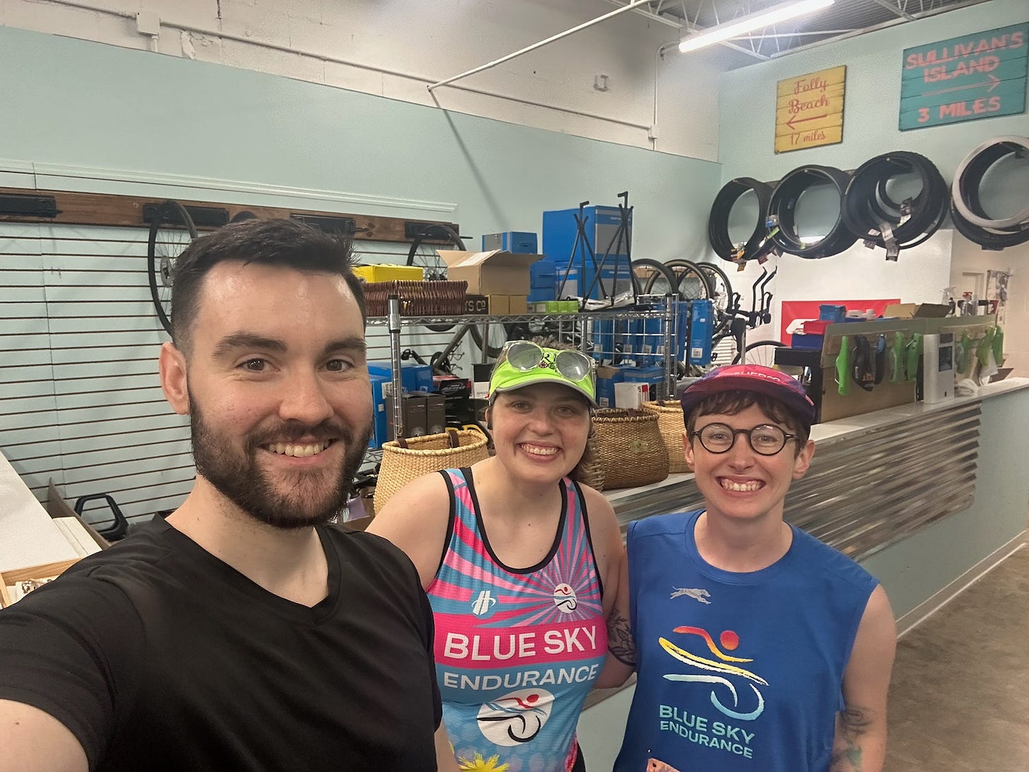 Dillon, Sydney (wearing blue and pink tank top and neon green visor), and Kimberly selfie in front of bike workshop at Blue Sky Endurance.