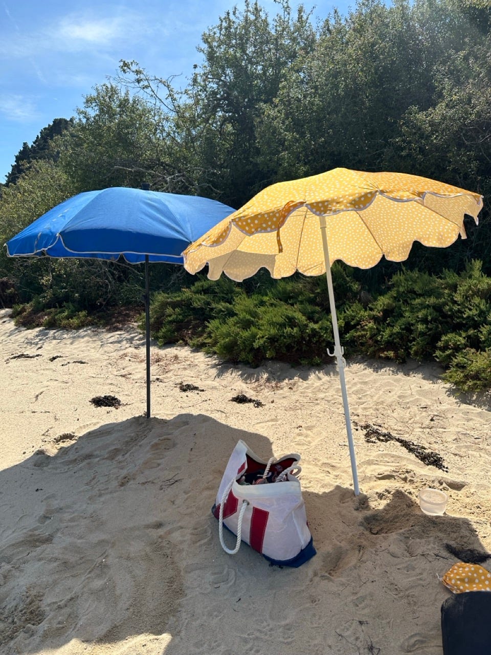 Two beach umbrellas — one yellow with white polka dots, one solid blue — on a quiet French beach, with a striped beach bag resting in the sand. Two beach umbrellas — one yellow with white polka dots, one solid blue — on a quiet French beach, with a striped beach bag resting in the sand.