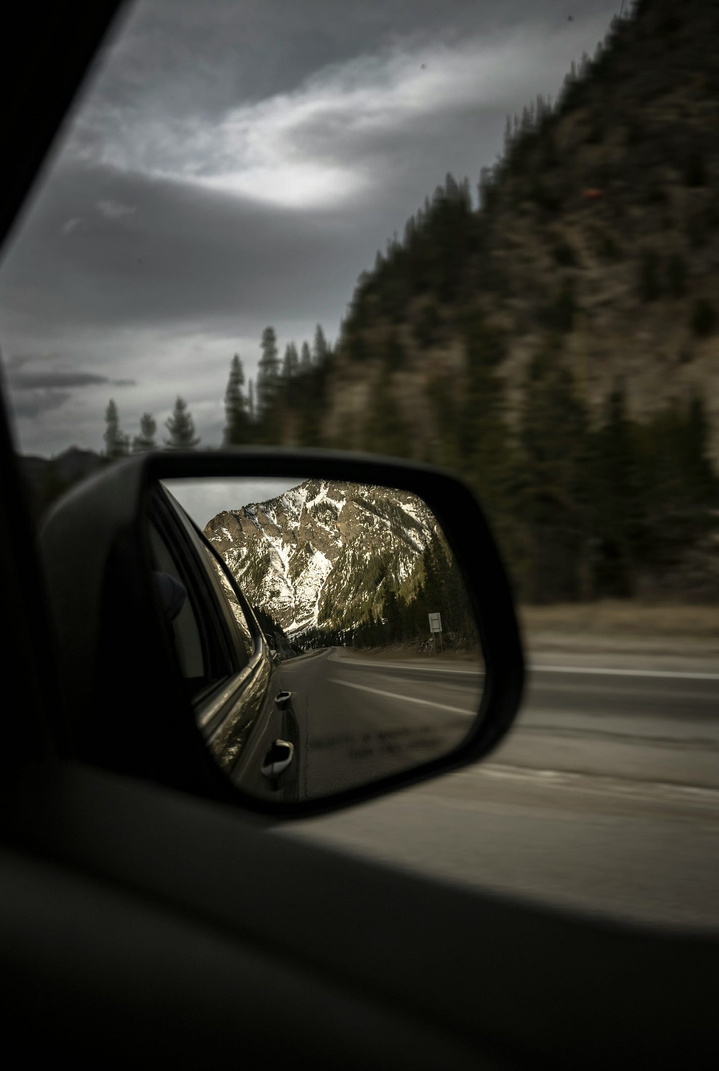 Snow-capped mountains in the side mirror of a car going down a tree-lined highway, against cloudy gray skies.