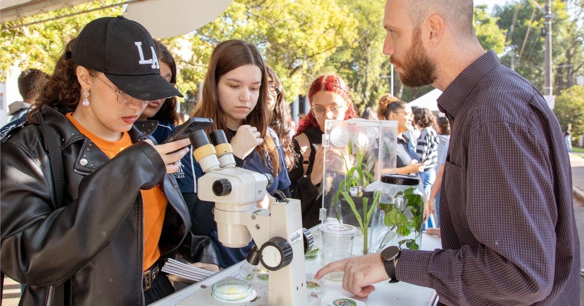 Estudantes em pé observam um microscópio numa mesa que também tem uma planta e do outro lado da emsa uma pessoa falando