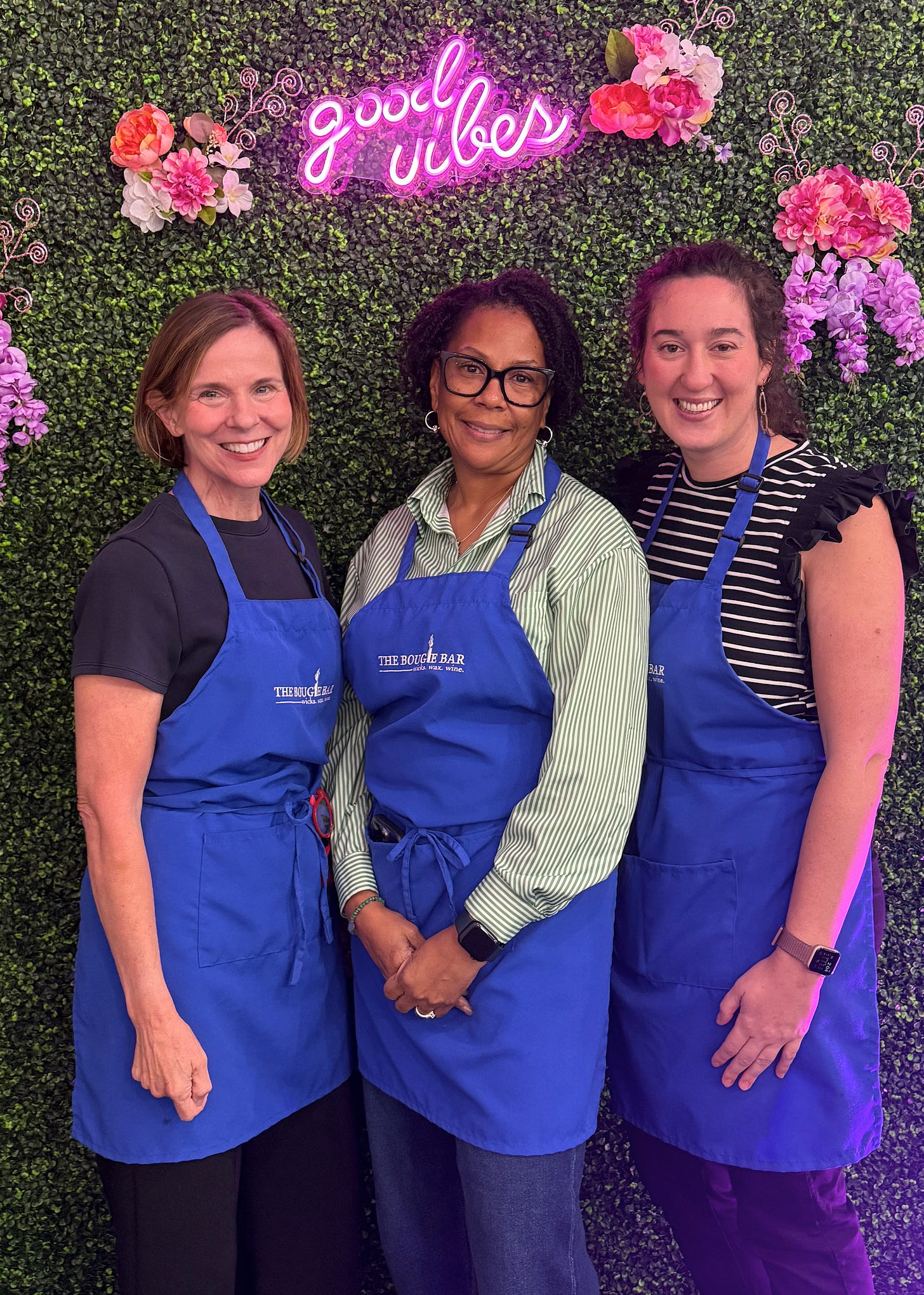 Three women wearing blue Bougie Bar aprons standing together in front of a floral wall with pink neon 'good vibes' sign