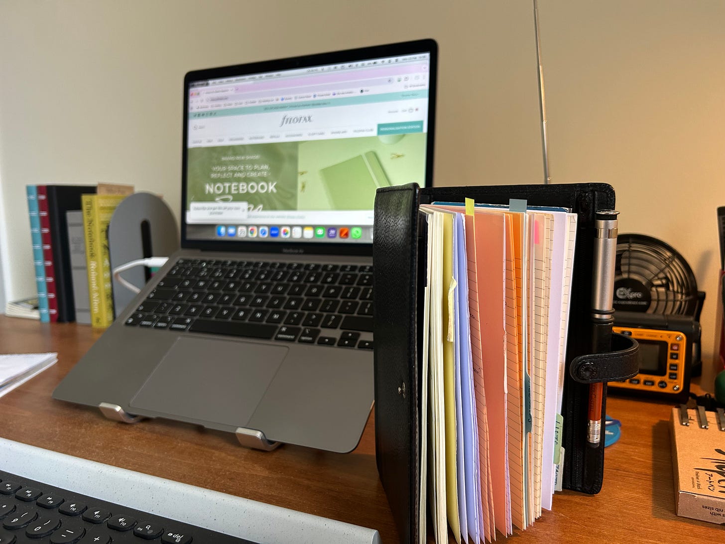 An office dsk with a personal Finsbury Filofax standing on end slightly open with an Apple Macbook Air laptop in the background with the Filofax website on the screen.