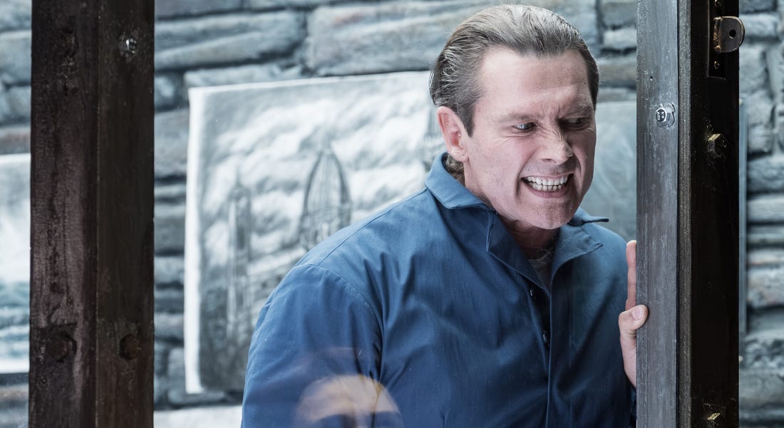A man in a blue jumpsuit snarling, with stone slabs and wooden beams behind him