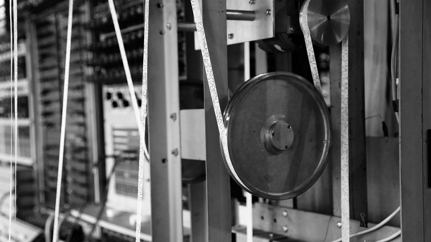 Photo of an early digital computer with a focus on punched paper tape passing around a wheel. Photo of an early digital computer with a focus on punched paper tape passing around a wheel.