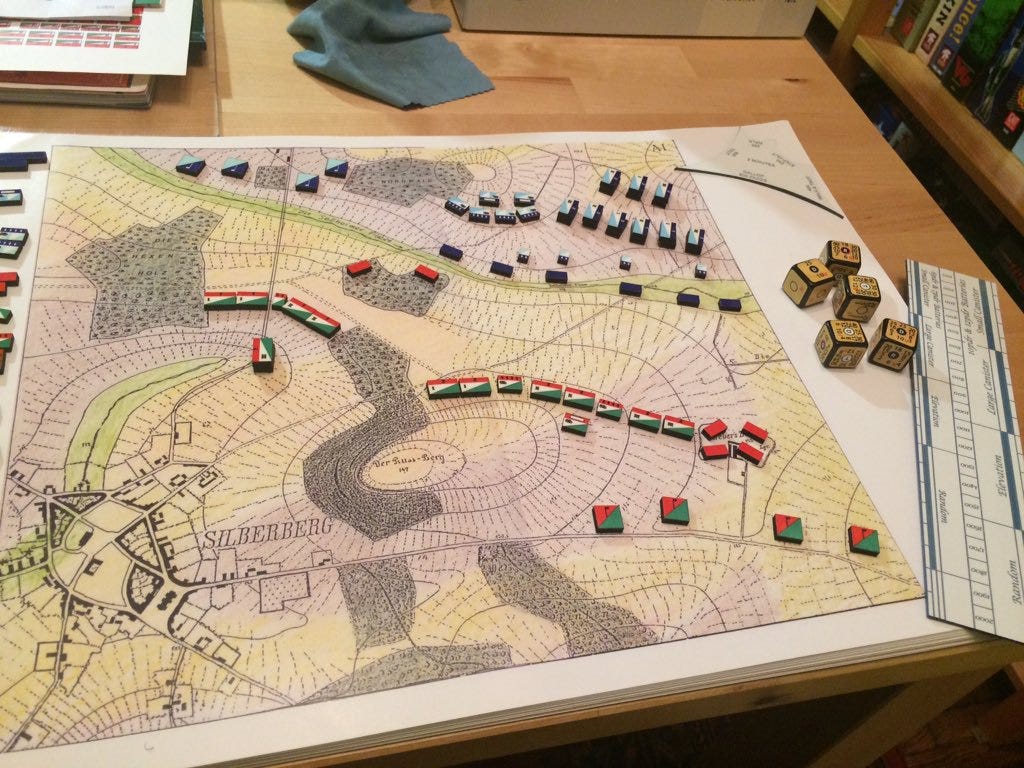 A wargame is laid out on a table. The map is covered in markers representing military units, and five dice rest on the right side of the map. A wargame is laid out on a table. The map is covered in markers representing military units, and five dice rest on the right side of the map.