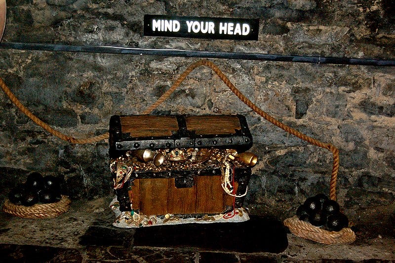 A wooden chest full of gold treasure in a stone basement. Cannonballs sit on coils of rope. A sign reads, "Mind your head."
