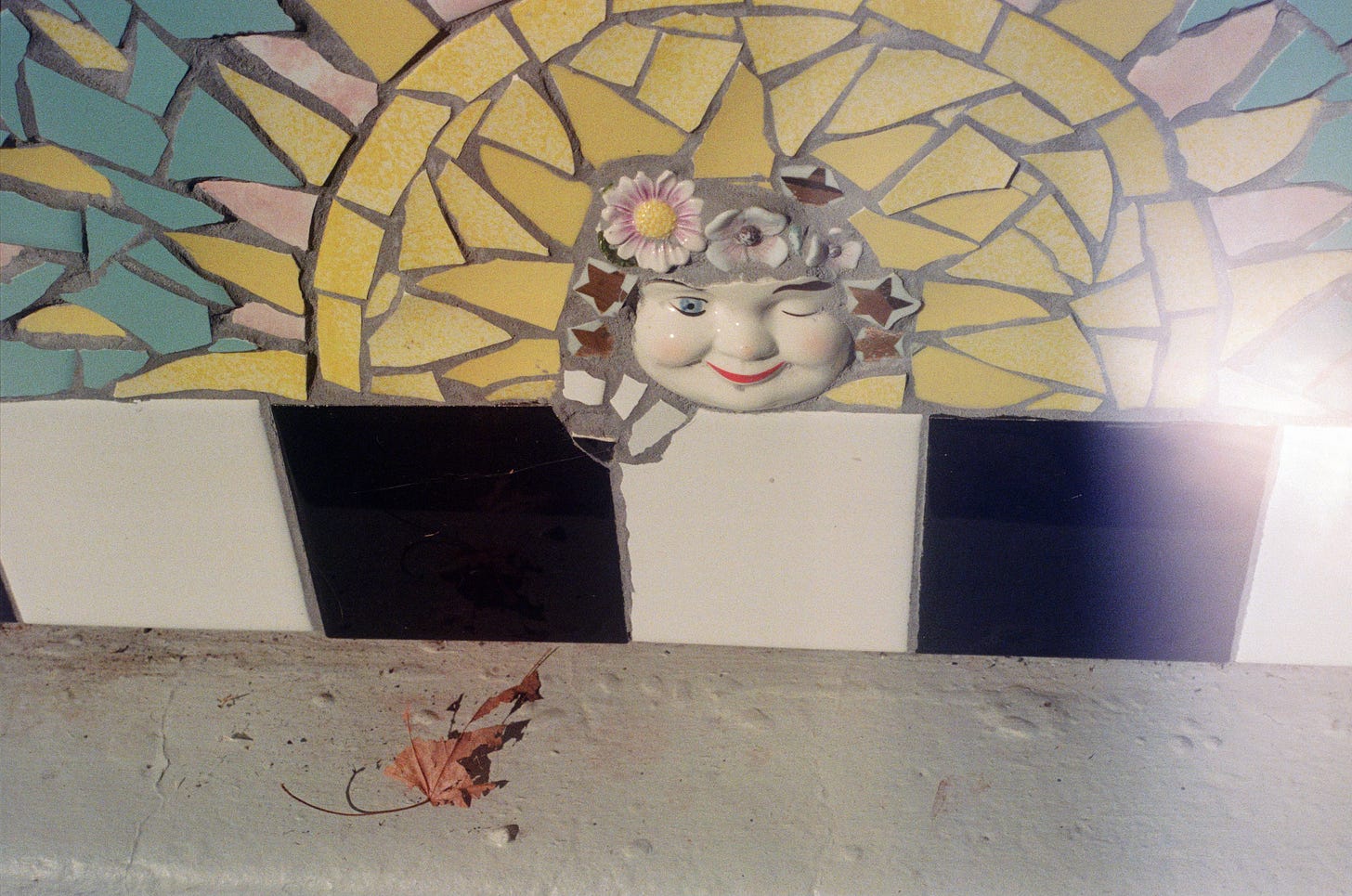 A film photograph of an outdoor wall shows colorful mosaic pieces that seem to create an image of a sun. In the center is a porcelain face, winking. A single brown leaf, crumbling, rests on the ground below. A light leak can be seen on the right side of the frame.