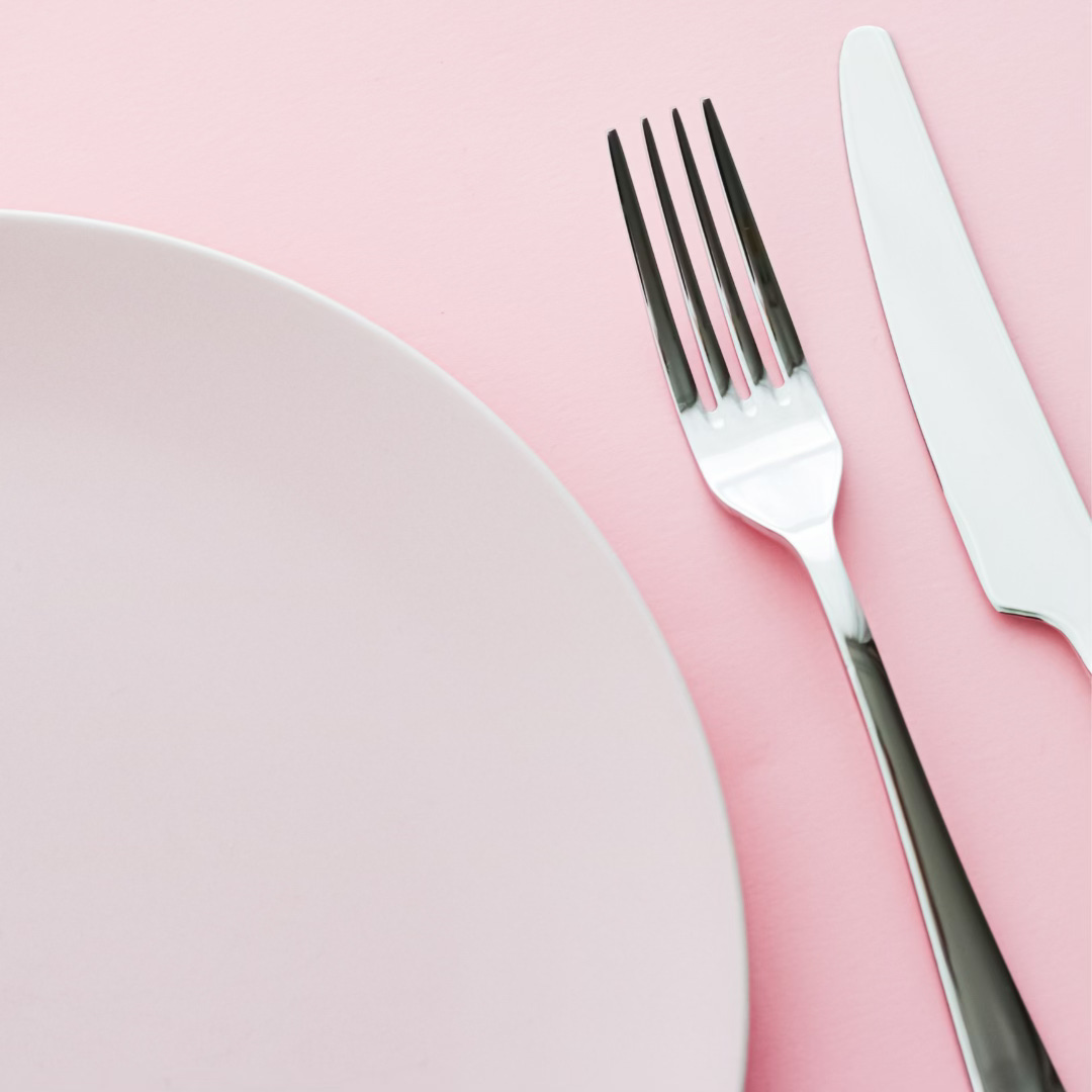 A silver fork and knife at an angle to a white plate. They are all on a pink background.