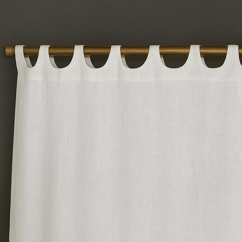 May include: White linen curtain panel with a scalloped top edge, hanging from a gold-colored rod. The fabric has a subtle texture, and the curtain hangs in soft folds. The background is a dark gray.