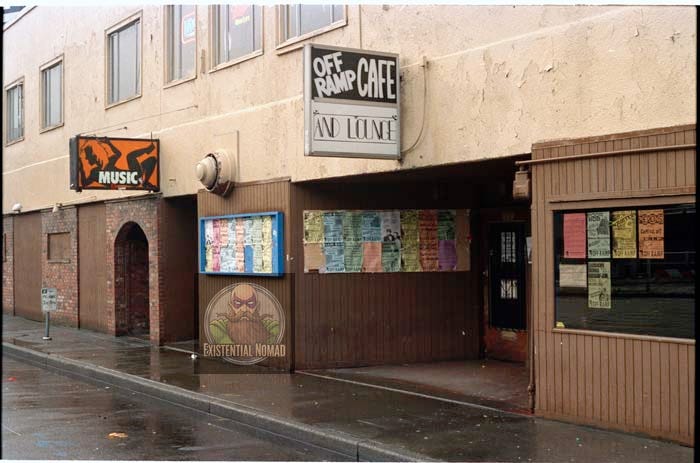  This is a photograph of the exterior of a music venue on a wet street. The building is beige, and the ground is slick with rain. There are two main signs on the building: one orange sign with the word "MUSIC" and a stylized logo, and another larger sign that reads "OFF RAMP CAFE AND LOUNGE." The entrance to the venue is recessed, and its exterior walls are covered in dark wooden paneling. The walls are covered in flyers for various bands.