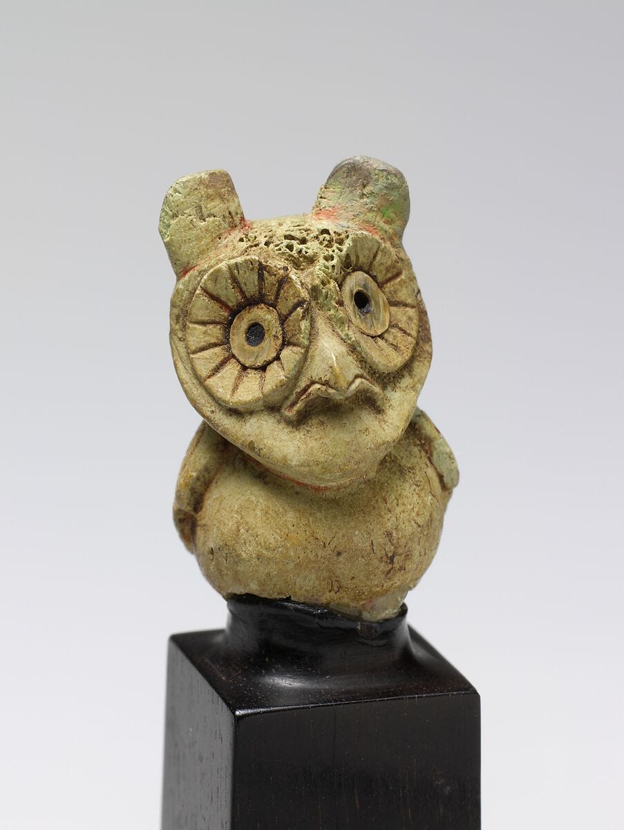 Small ancient ceramic figurine of an owl, squat and round-bodied with large circular eyes and small ear tufts, in a weathered greenish-tan glaze with traces of red. Mounted on a small black pedestal.