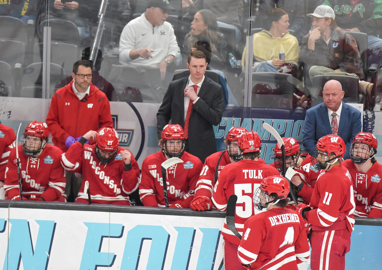 Nick Oliver wearing a suit and tie standing behind the bench of players. Oliver is flanked by a Badgers athletic trainer and UW head coach Mike Hastings
