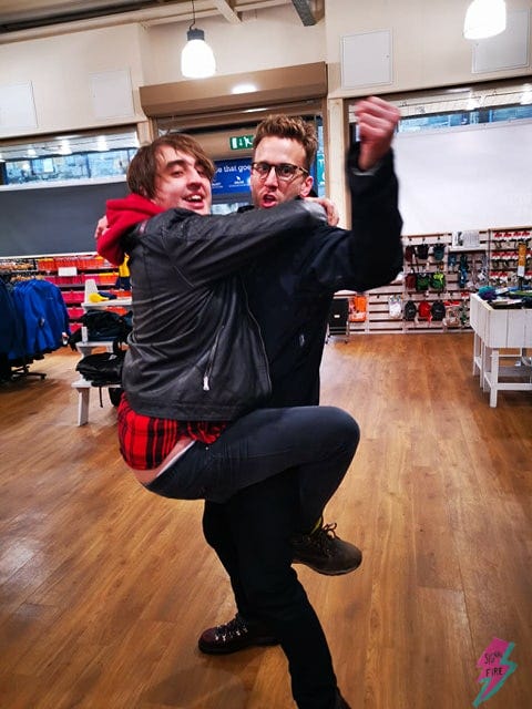 Ash Raymond James and Tyler Knott Gregson dancing in a Scottish outdoors shop