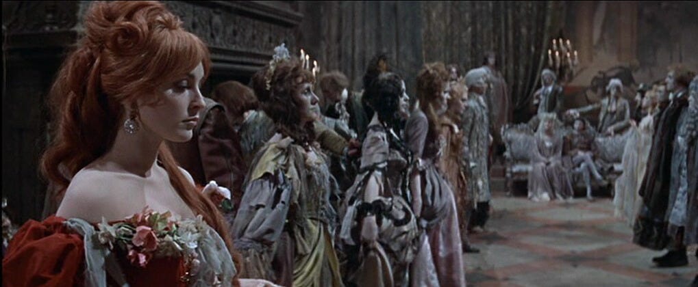 quatro mulheres em vestidos suntuosos de época enfileiradas lateralmente, com pessoas atrás e algumas ao fundo, todos com roupas que parecem de gala e cabelos bem arrumados. A mulher mais próxima da câmera é ruiva, branca, com um vestido vermelho