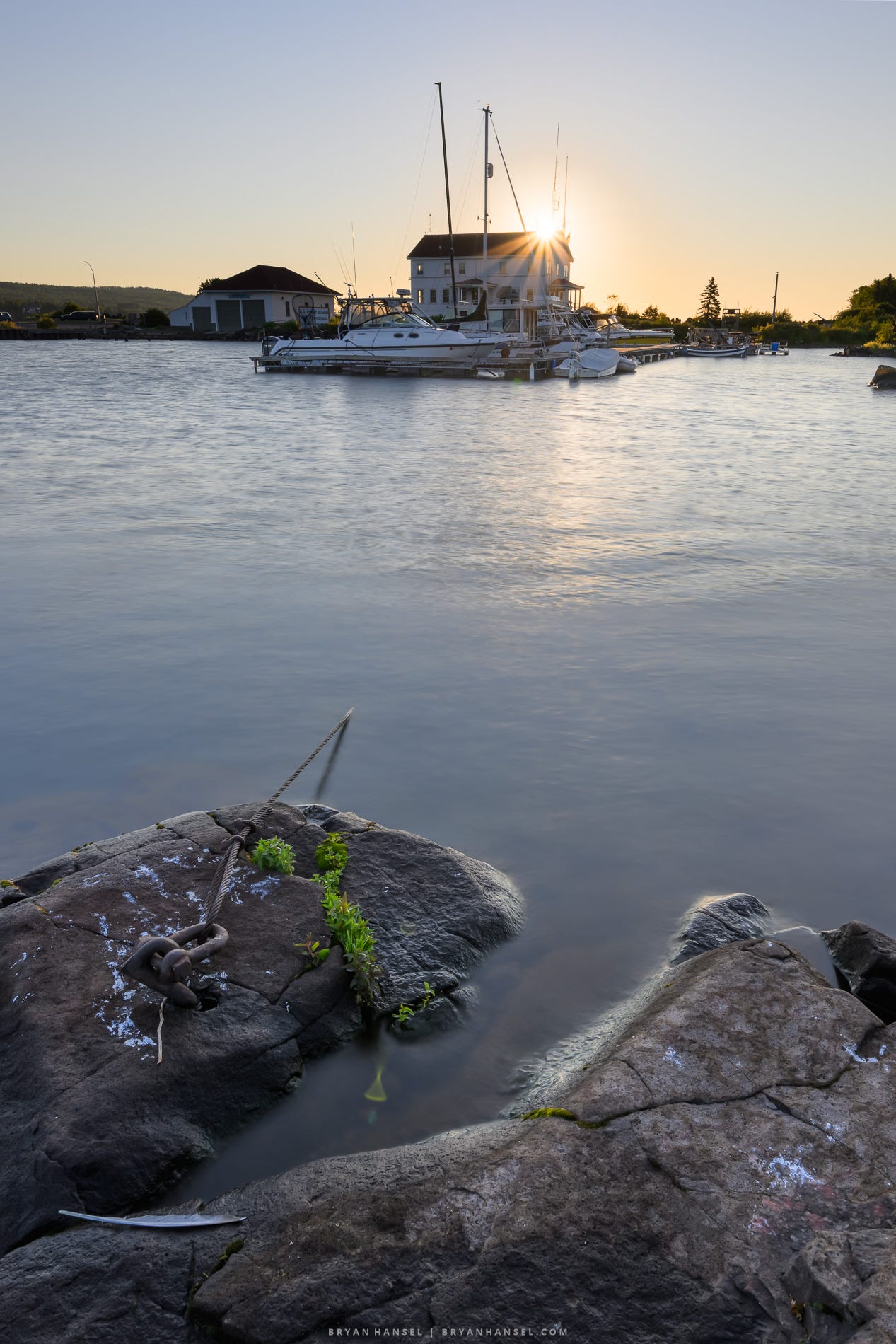 Sunrise over the Grand Marais harbor, with boats moored in front of the Coast Guard building. Smooth water reflects the warm morning light, while granite shoreline rocks and a mooring chain fill the foreground. HDR version