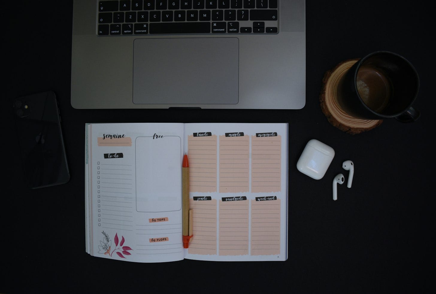 Overhead view of a dark desk with a laptop, an open weekly planner and pen, a phone, wireless earbuds and a mug of coffee. Overhead view of a dark desk with a laptop, an open weekly planner and pen, a phone, wireless earbuds and a mug of coffee.