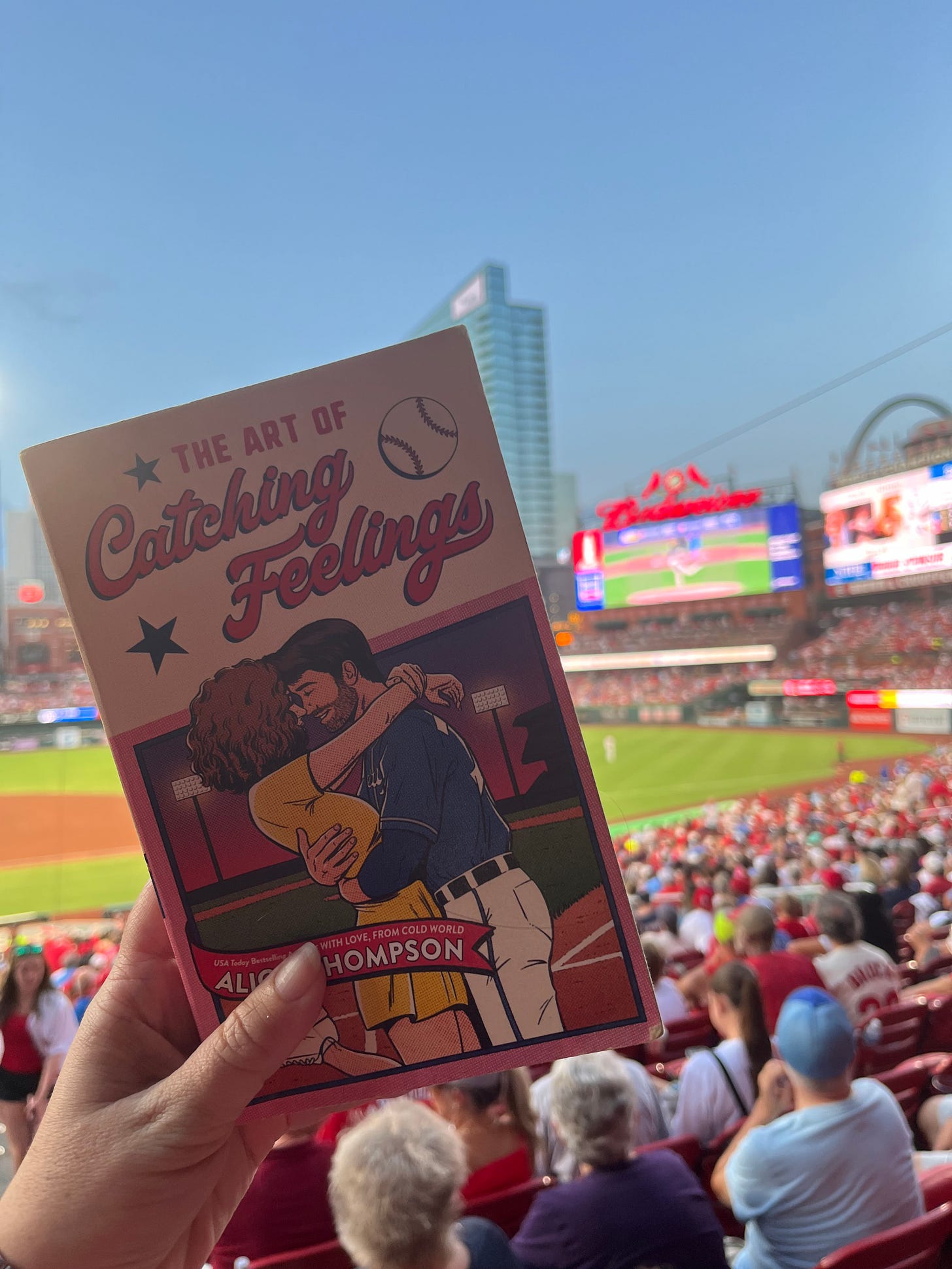 Photo of my hand holding up copy of THE ART OF CATCHING FEELINGS in front of field at Busch Stadium, featuring the jumbotron and the Arch in the background