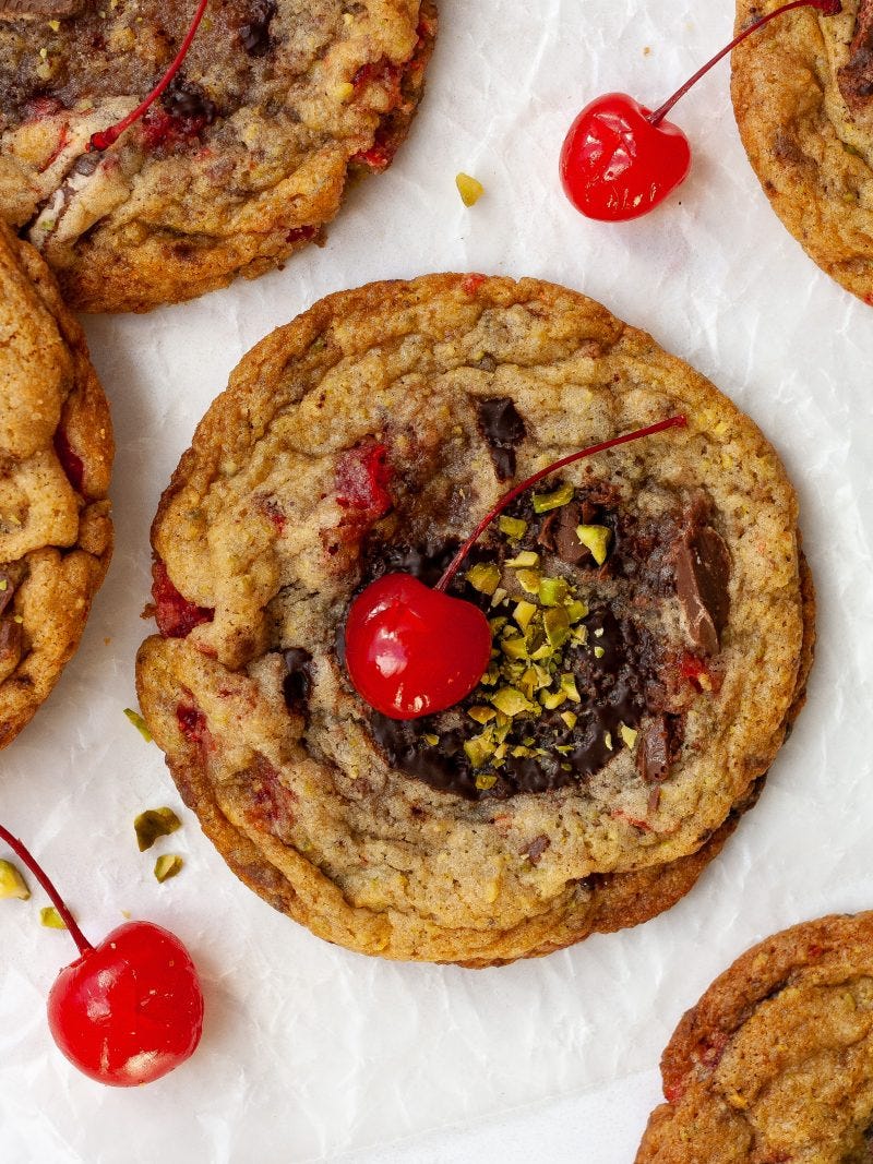 brown cookies with chocolate chunks, sprinkles of chopped green pistachio, and a red maraschino cherry sit on a white surface