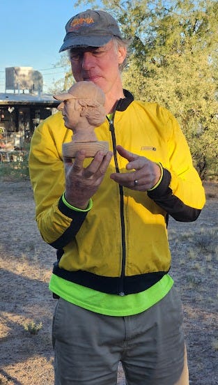 A white man in a yellow jacket looks at a bust of his head made from brown clay.