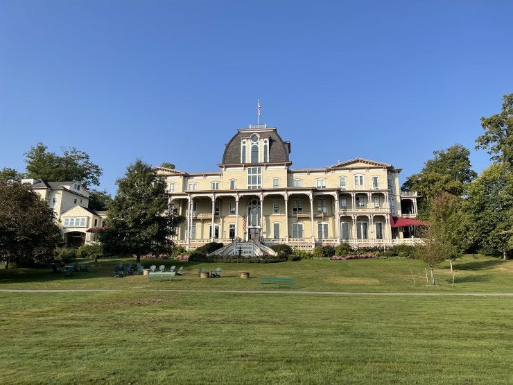 The Athenaeum Hotel at Chautauqua Institution, a yellow Victorian hotel with a distinctive dark mansard roof, central tower, and multi-tiered wraparound porches with white columns. The historic building sits on a manicured lawn furnished with Adirondack chairs and is surrounded by mature trees under a clear blue sky.