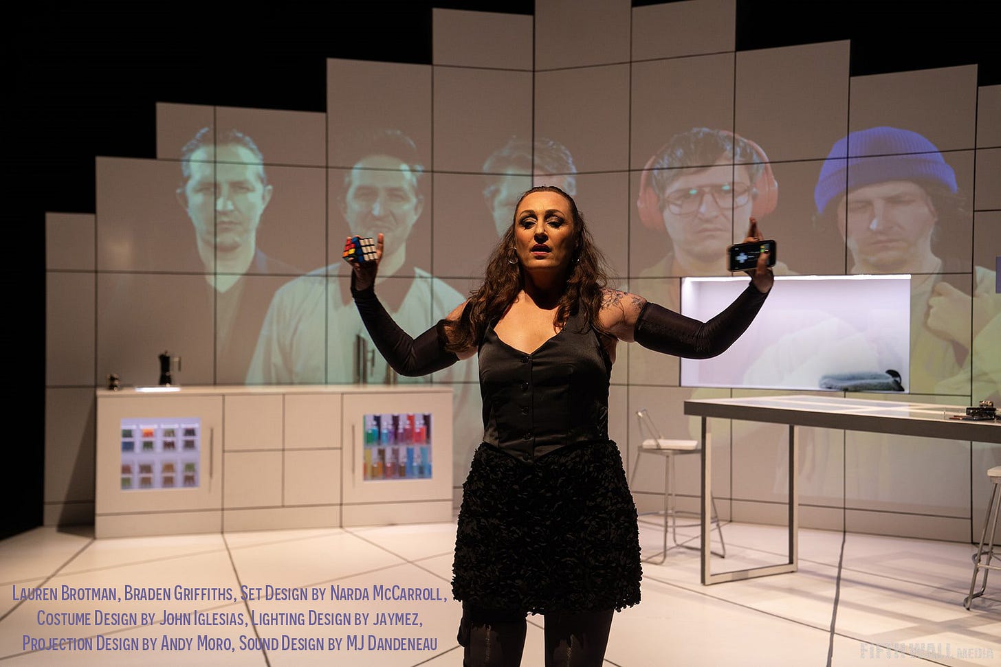 A woman stands on stage with her arms outstretched. She is holding a Rubix Cube in one hand and a cellphone in her other. There are projections on the wall behind her of five men, all played by the same actor.