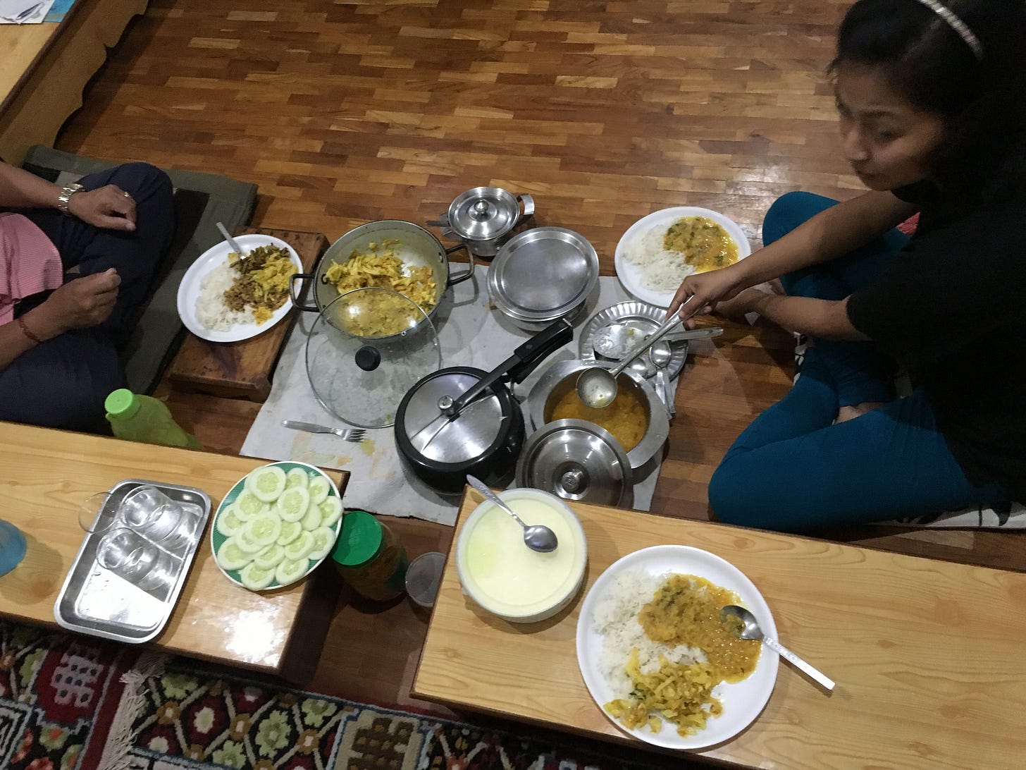The bottom half of a seated man and the side view of a brown woman wearing a black top and blue pants. Pots, pans, and plates are placed on wooden floors and tables, holding dishes such as dal, cooked cabbage, cucumbers, and rice.