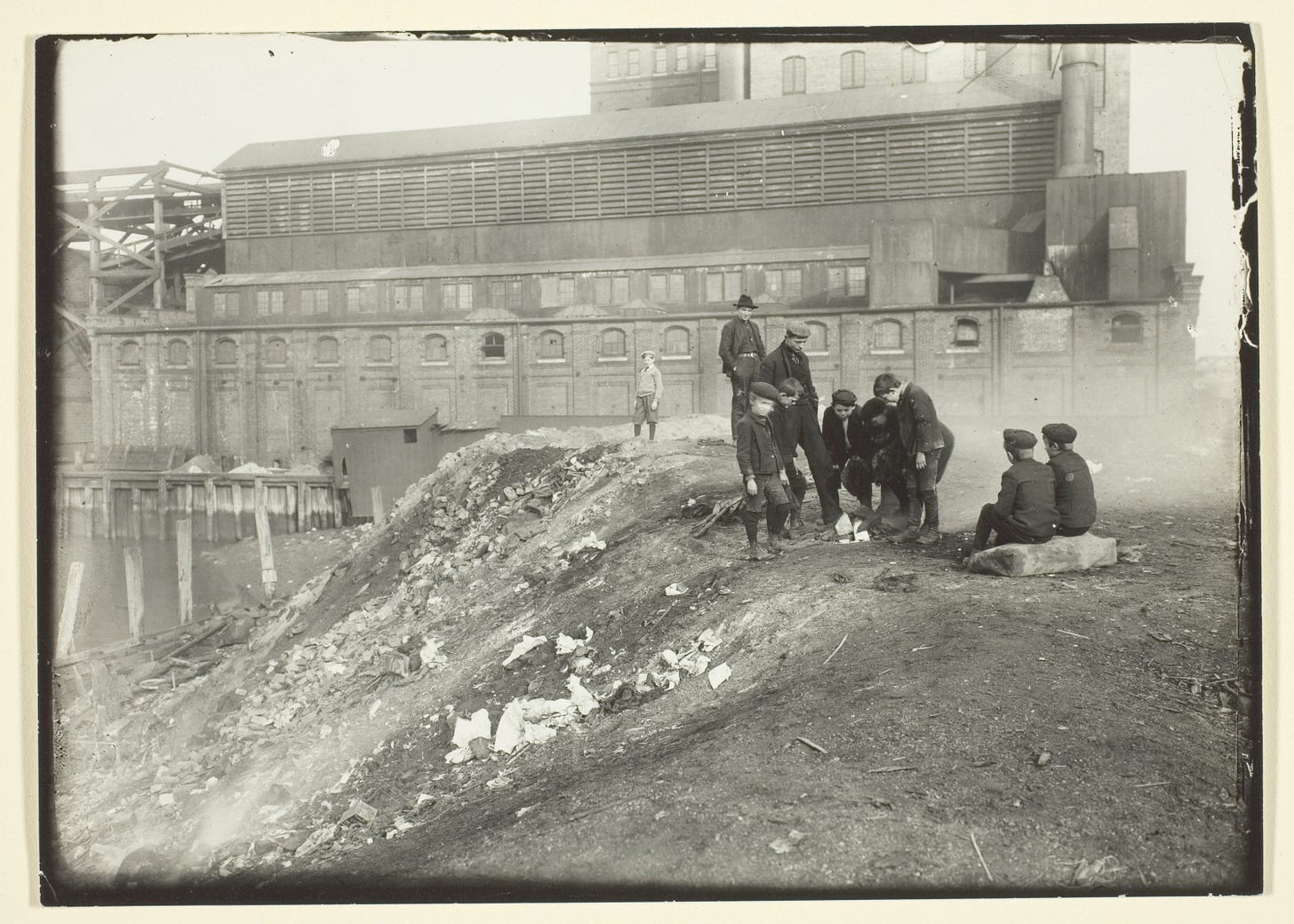 Black and white gelatin silver print from 1906. There is a large industrial building to the right of a wooden bridge in the background. In the foreground are a group of young boys who appear to be looking at something on the ground and making plans. An older man in a light colored sweater stands a bit off from the group of boys, watching them.
