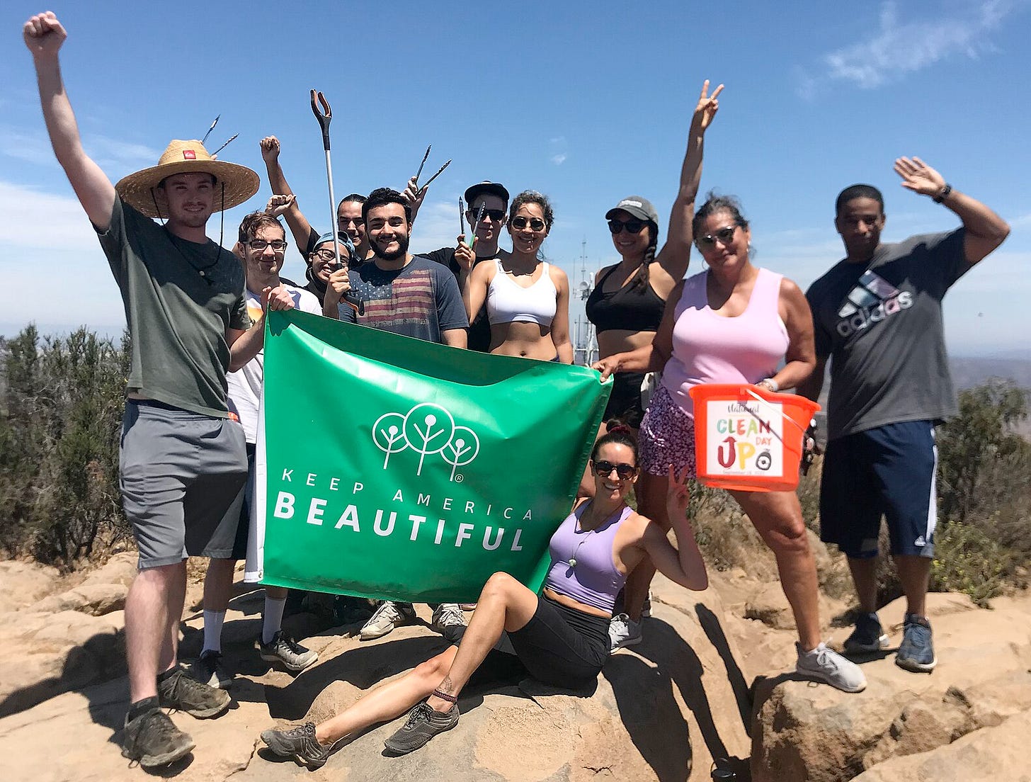 a group of young people in summer clothes standing on a rocky sandy outcrop on top of a bluff looking over the ocean, holding up a green "keep america beautiful" banner and cheering