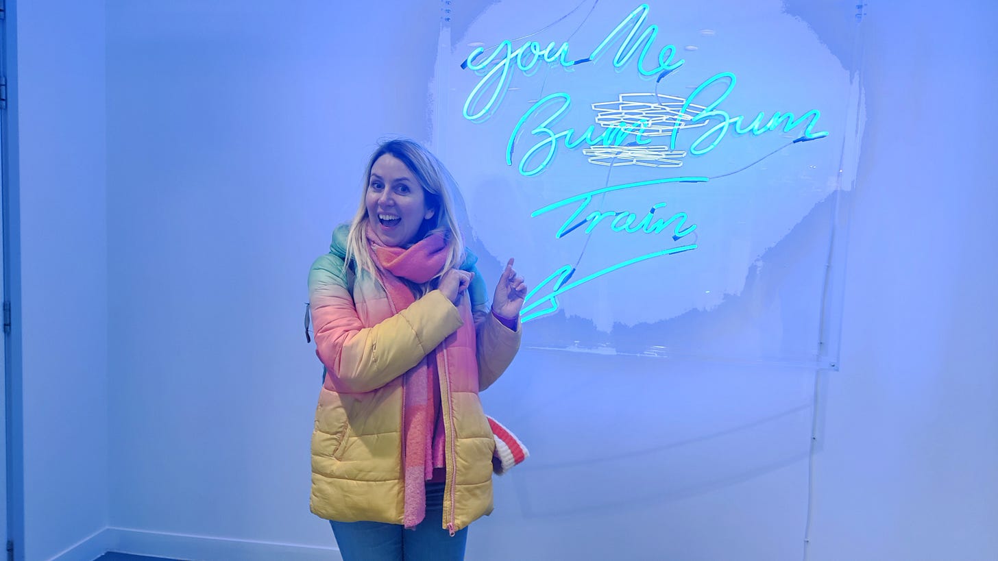 A photo showing Emily Jupp by a neon sign reading you me bum bum train