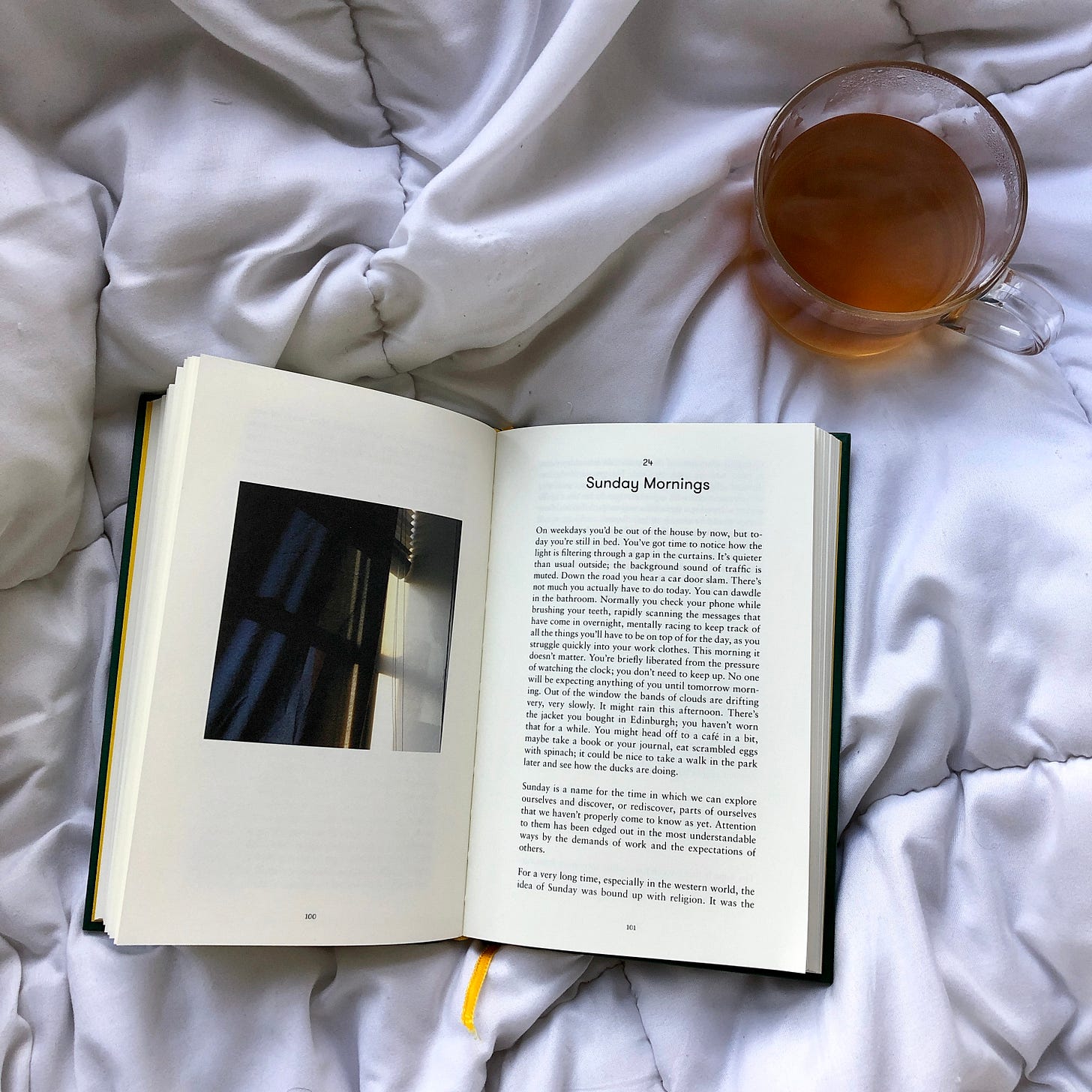 An overhead view of a book opened to a chapter called "Sunday Morning” on top of a bed. There is a cup of tea next to it.
